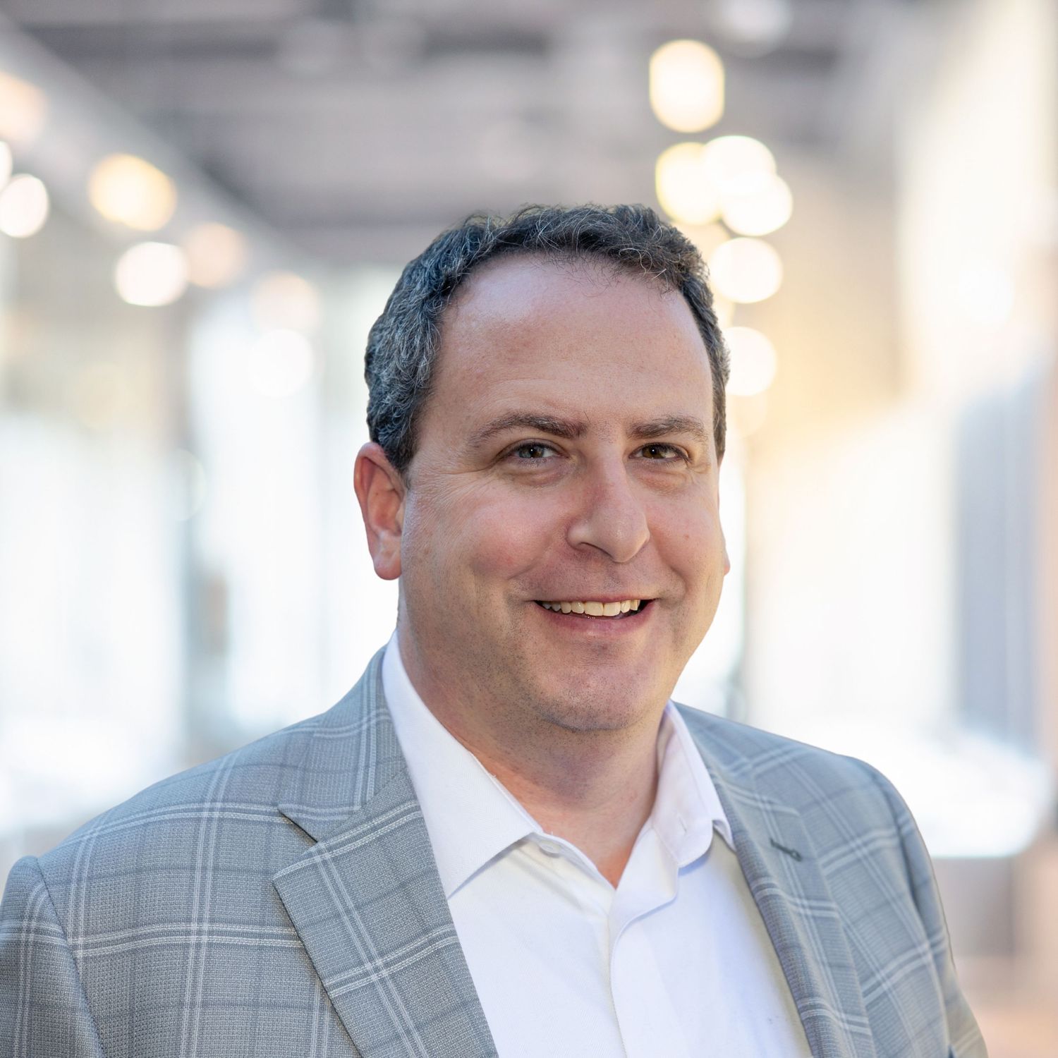 Professional headshot of someone in a gray plaid blazer and white dress shirt smiling in an office setting.