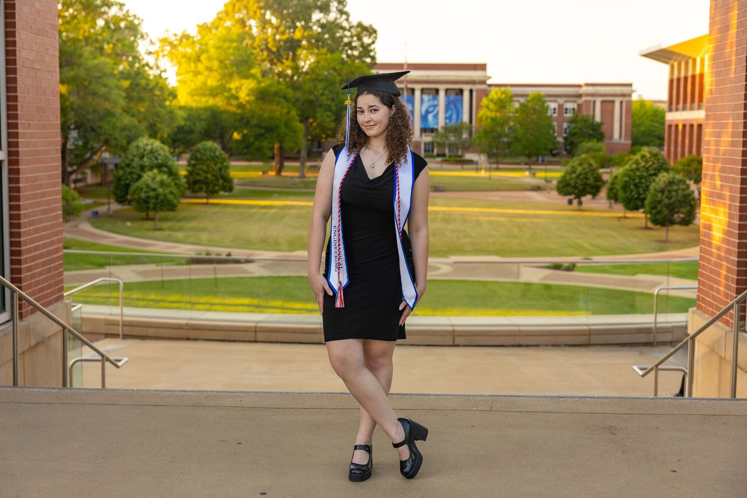 Young woman in graduation cap and gown poses outdoors on a sunny college campus in a black dress.