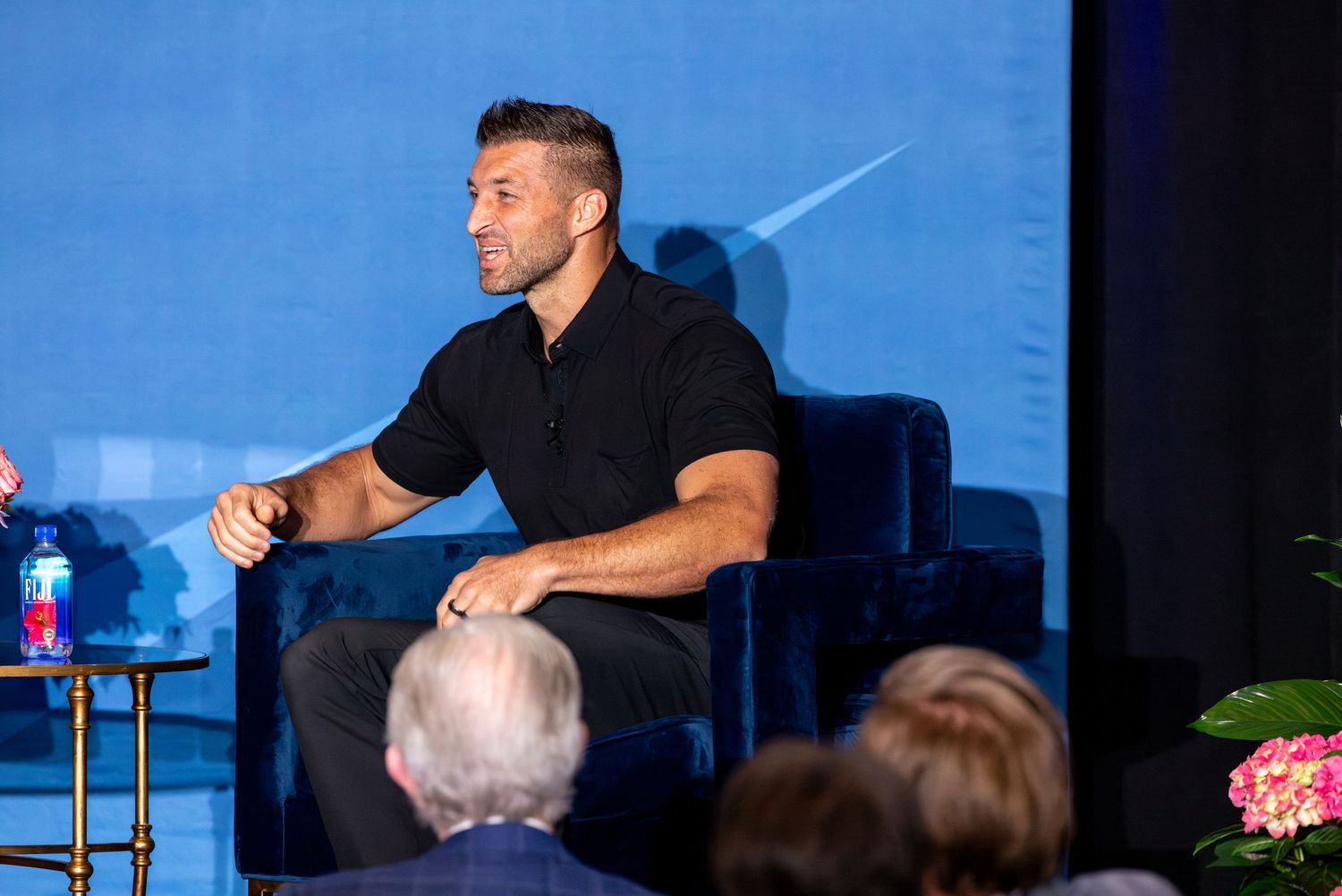 Man in black shirt sits on blue velvet chair on stage during a panel discussion event with audience.
