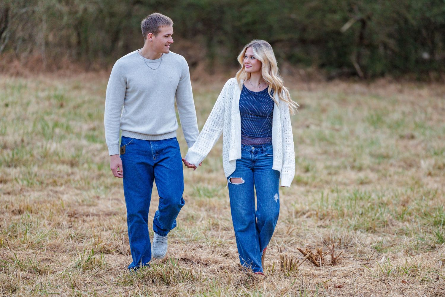 A couple in blue jeans and light tops walking together in a grassy outdoor field.