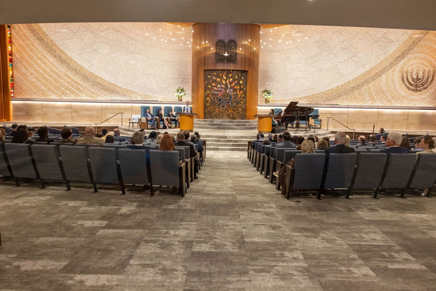 Elegant synagogue sanctuary with rows of chairs facing a decorative bimah stage with grand piano and menorah.
