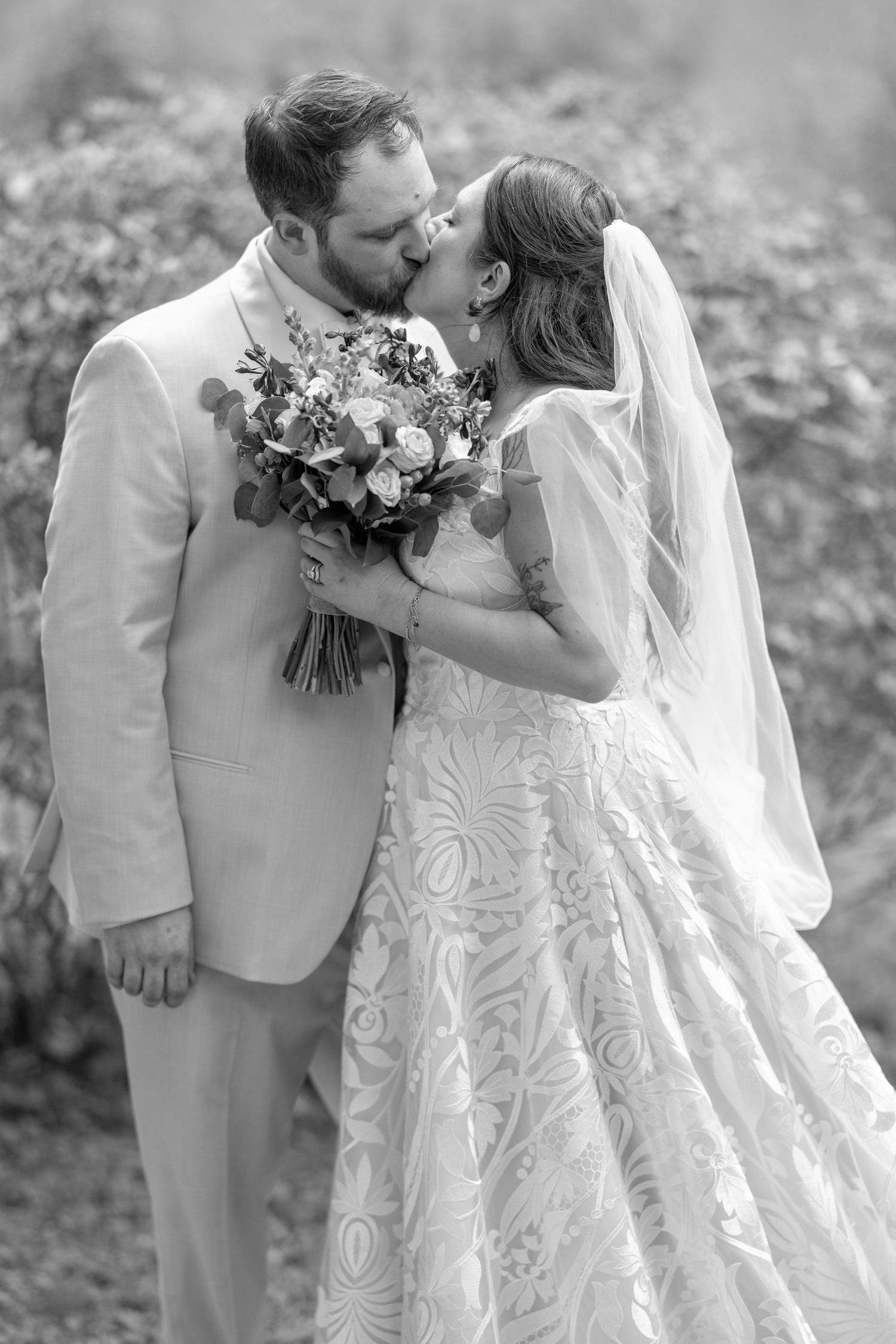 Black and white photo of a bride and groom sharing a kiss, bride holding a floral bouquet in a lace wedding gown.