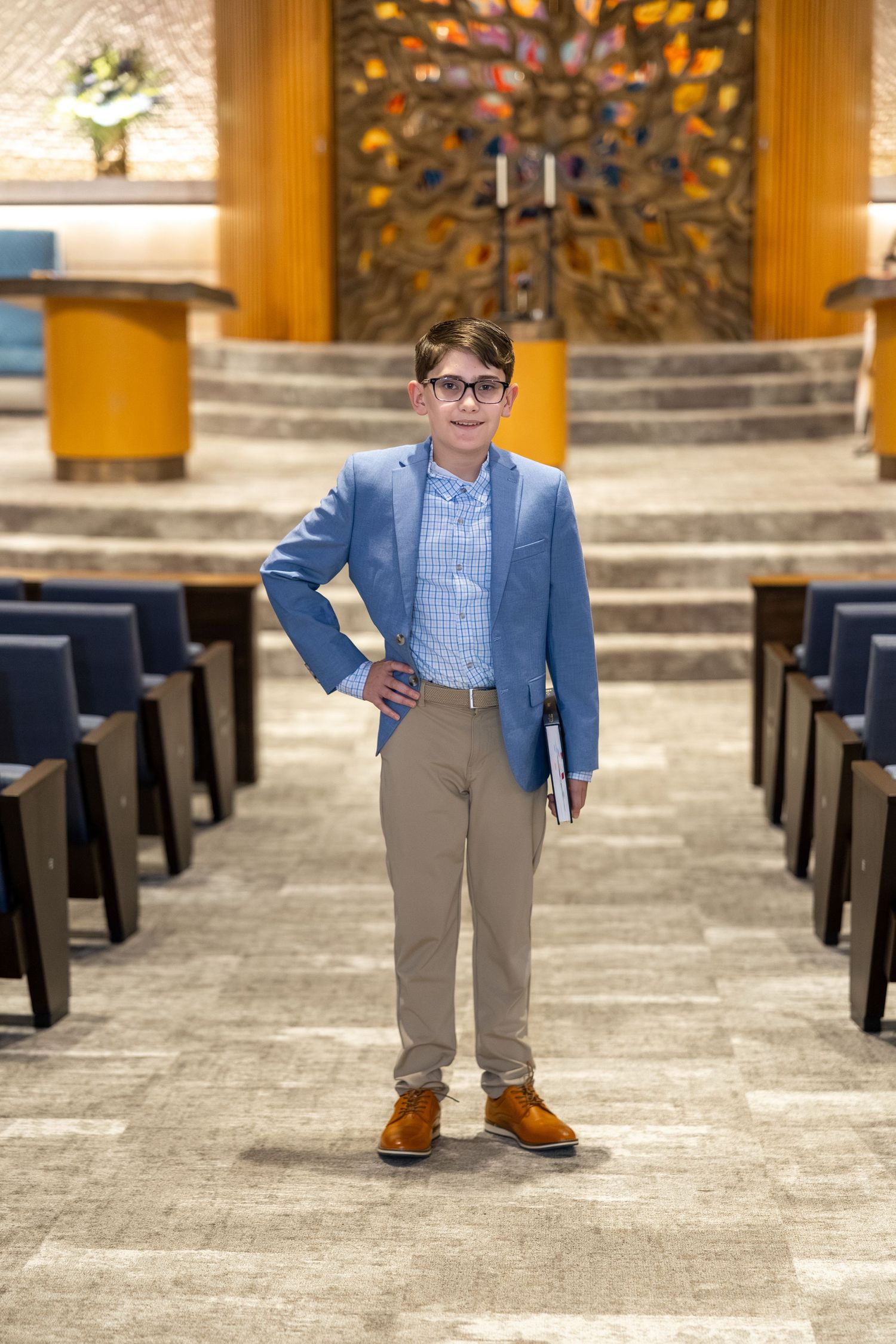 Young boy in blue blazer and khakis stands confidently in synagogue aisle holding a book at his Bar Mitzvah.
