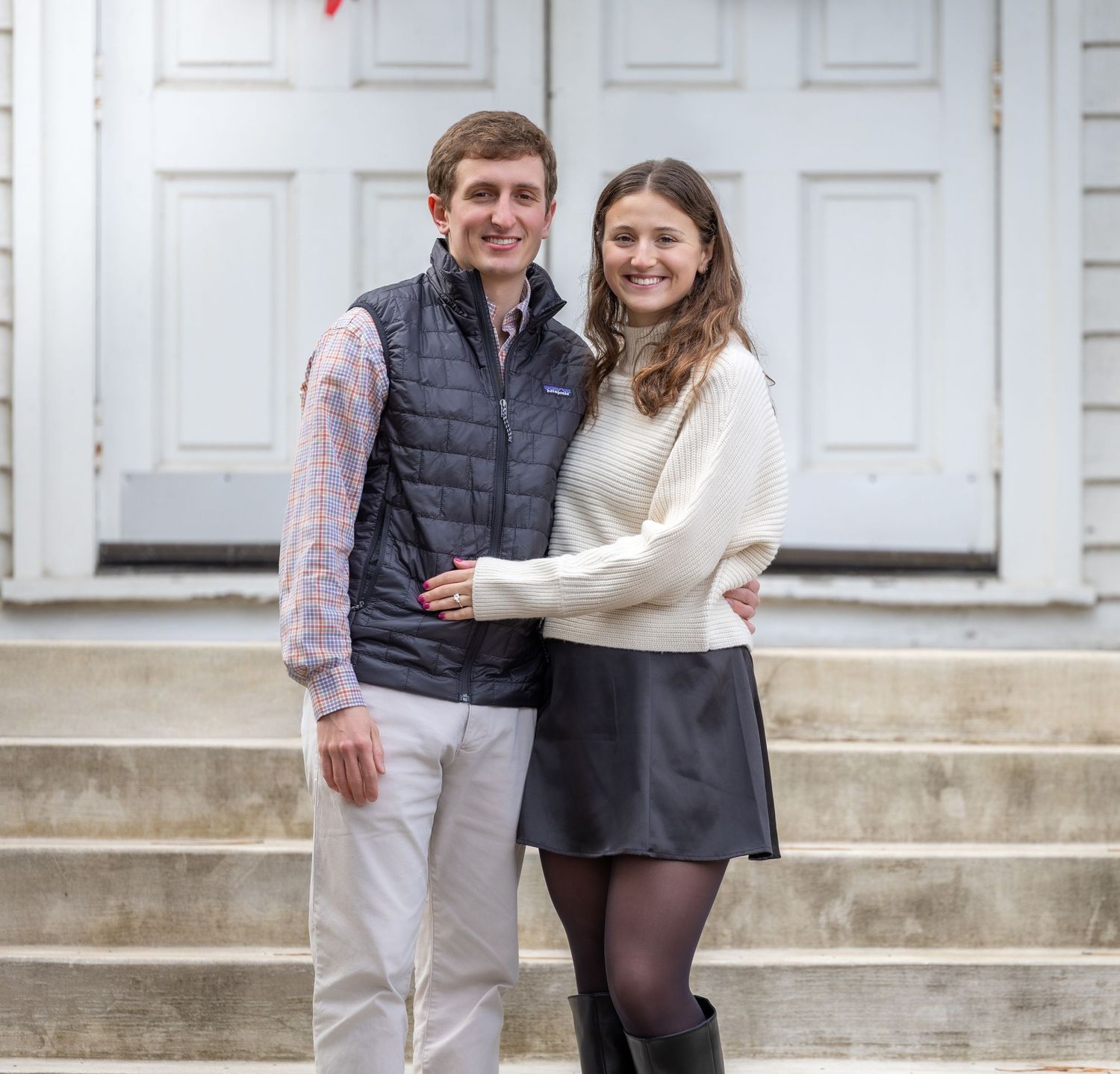 A couple stands together smiling on white stone steps outside a building with classic white doors.