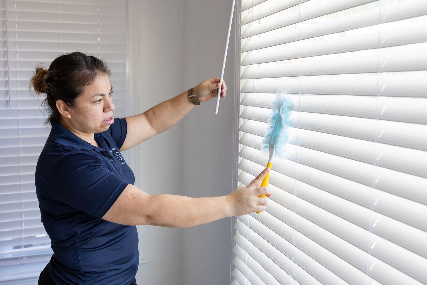 Cleaning service worker dusting white window blinds in home.