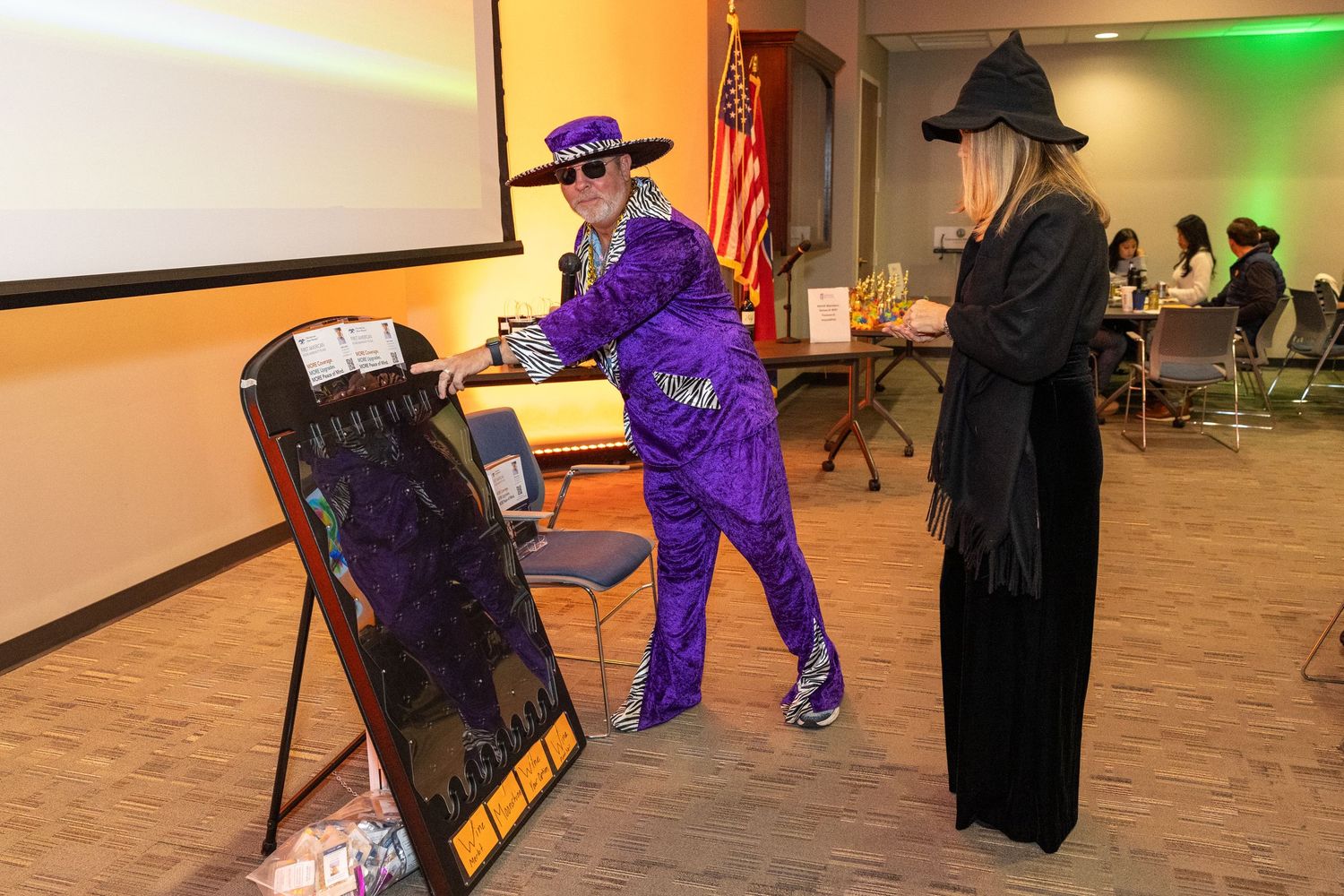 Halloween party attendees in costumes mingling in a decorated room with green and orange lighting.
