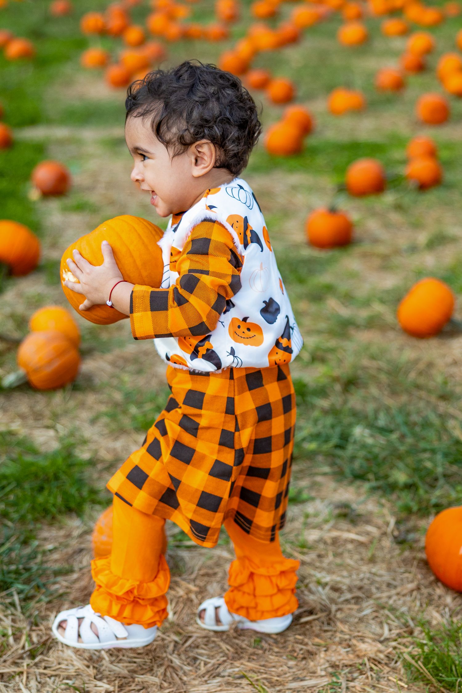 Small child in pumpkin-themed outfit picks up orange pumpkin from grassy field.