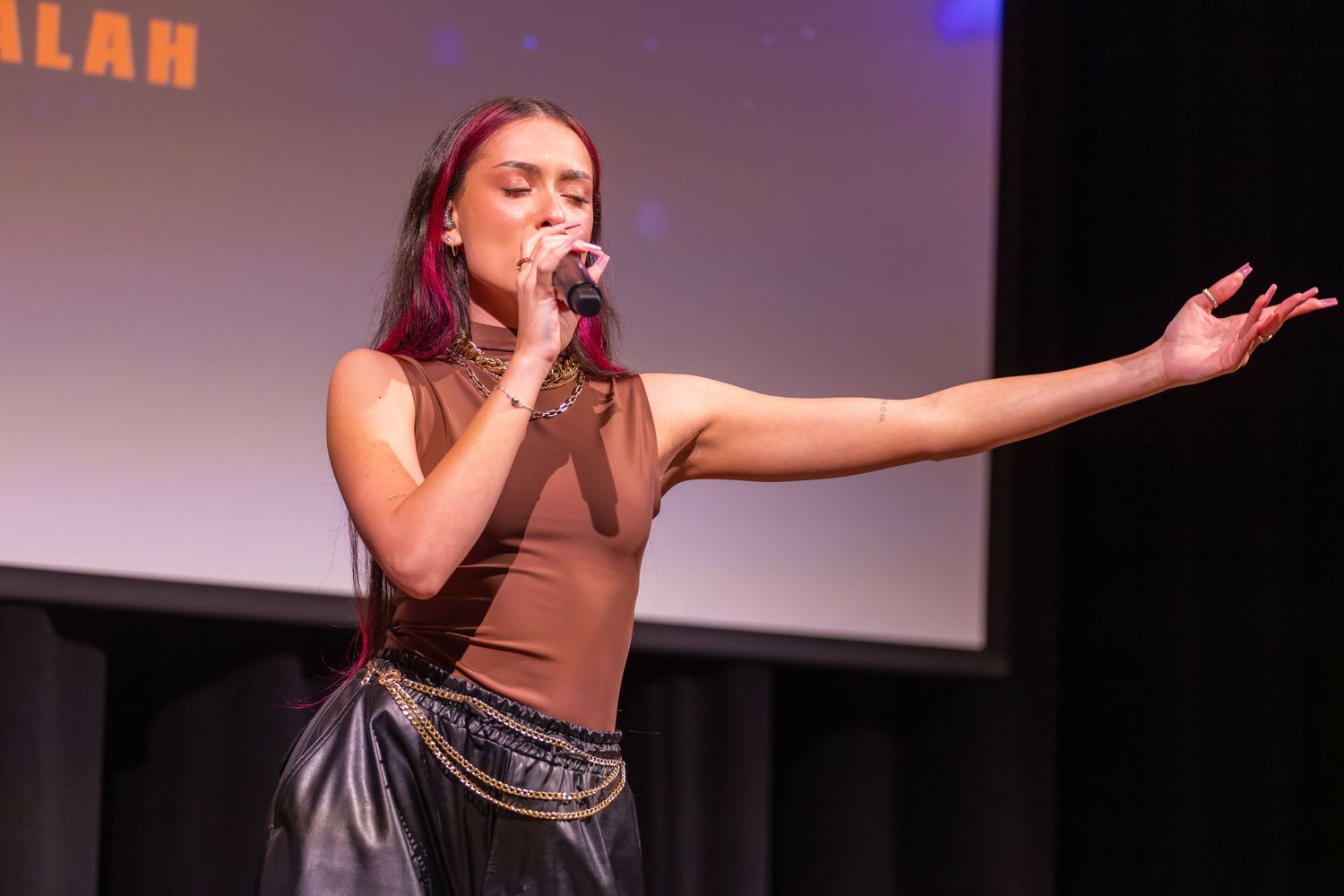 Performer in brown top and dark skirt sings passionately on stage with purple lighting.