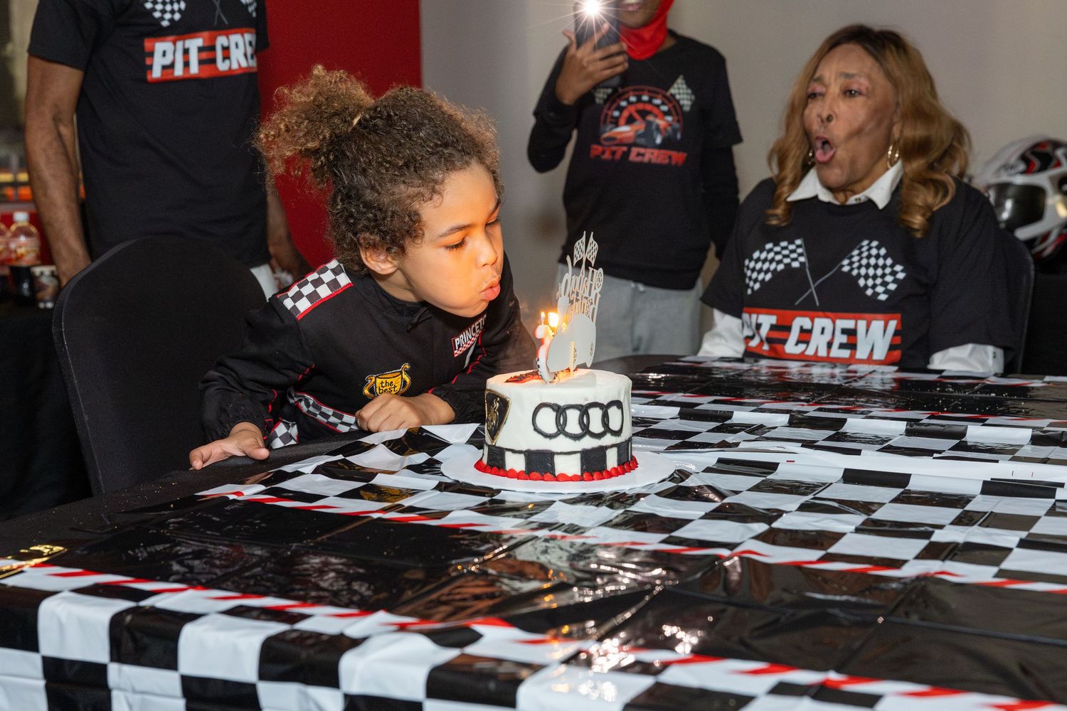 A young person blows out birthday candles on a racing-themed cake with checkered decorations.