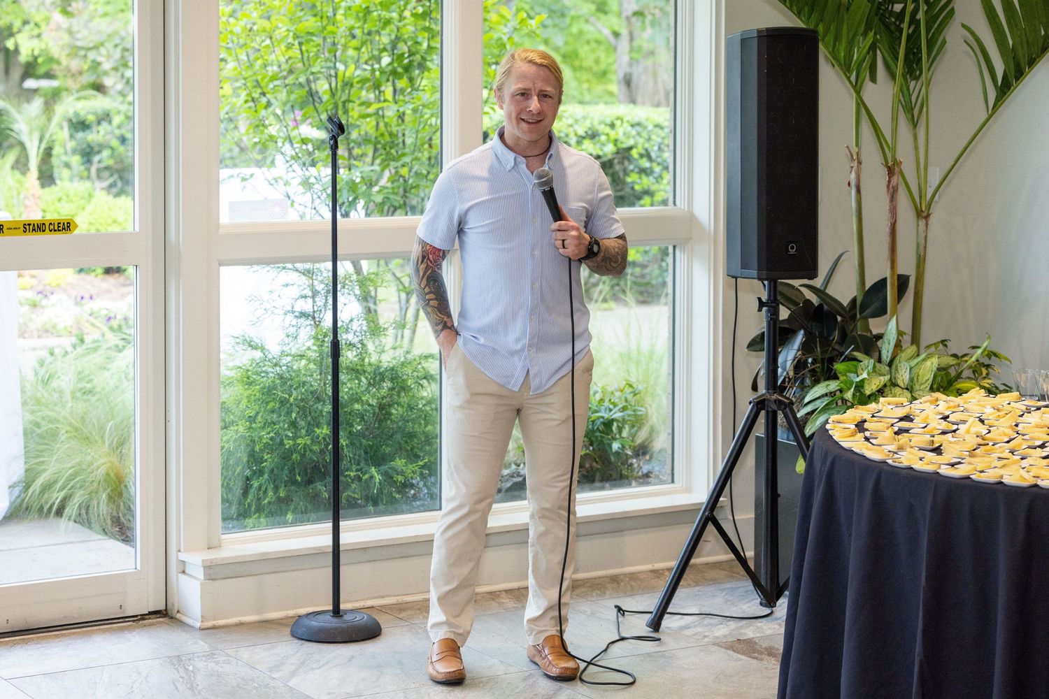 Man in white shirt holds microphone on stand at indoor event with speaker and tropical plants.