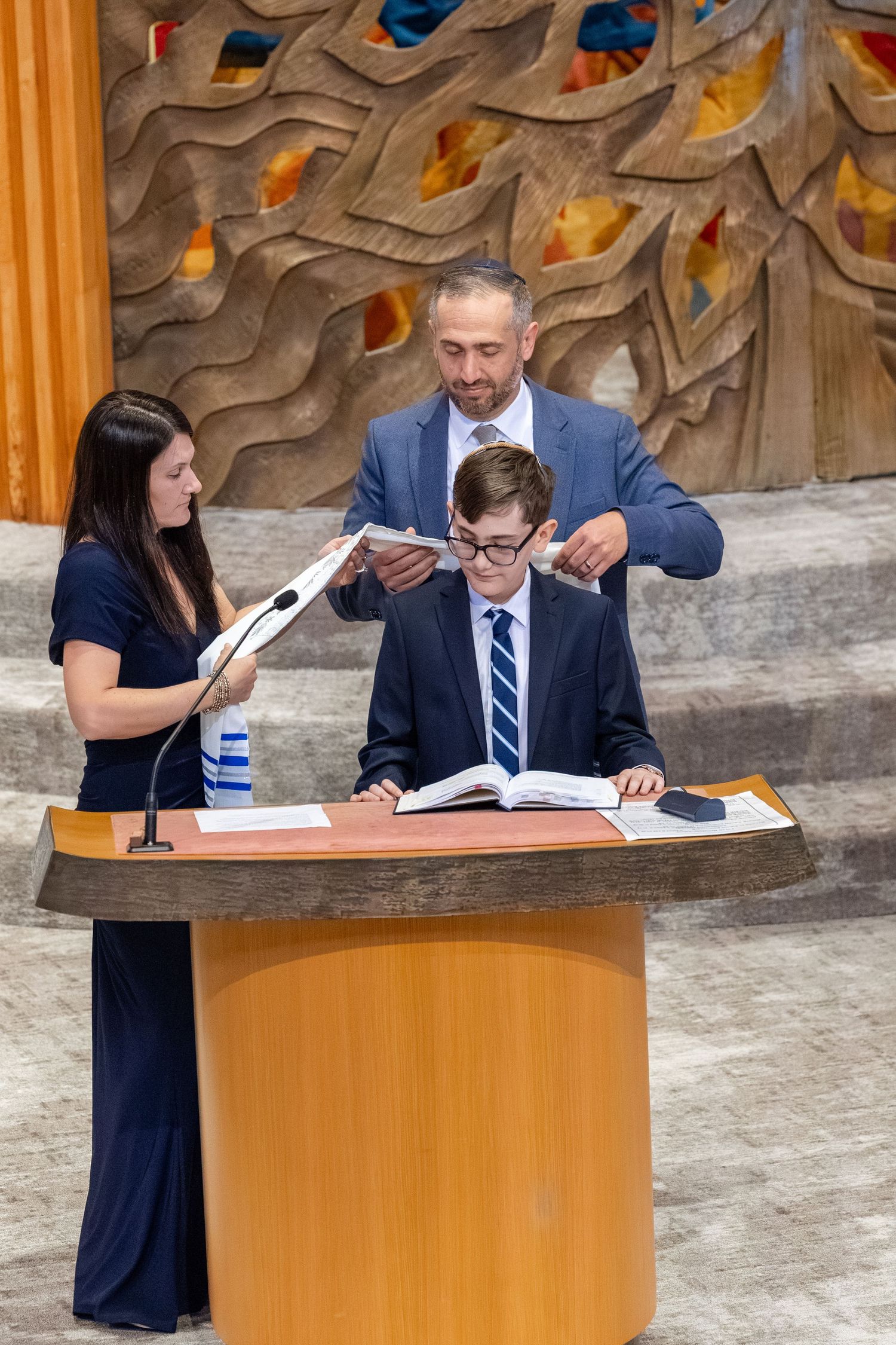 Boy reading at podium during Bar Mitzvah ceremony with parents placing tallit on his shoulders.