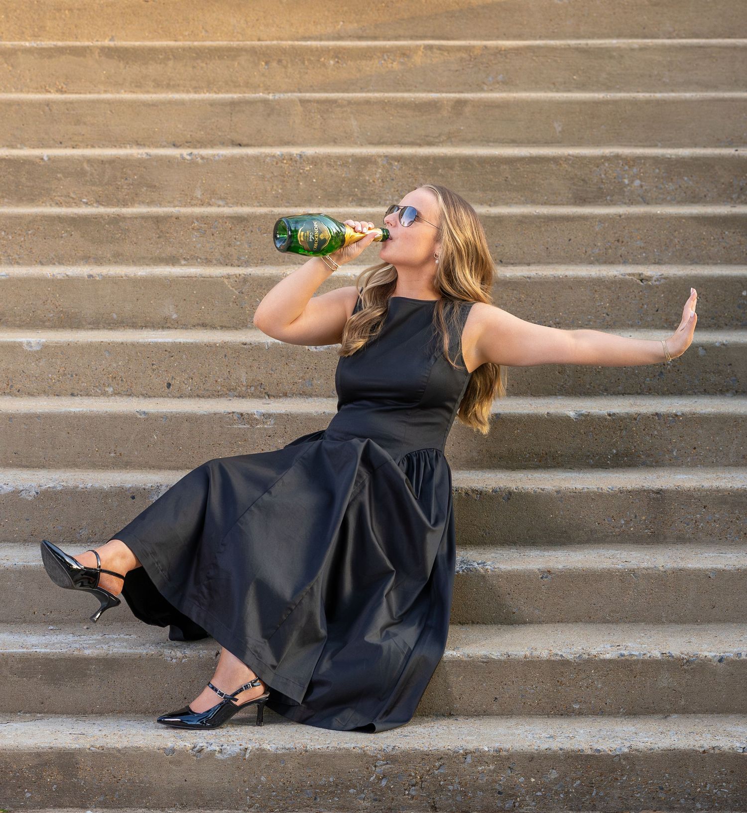 Woman in black dress drinking from a green bottle while sitting on stone steps outdoors.