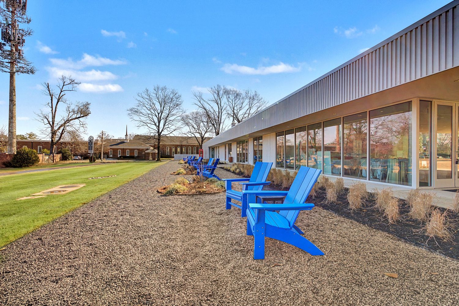 Blue Adirondack chairs line a gravel patio beside a modern building with large windows and a lush green lawn.