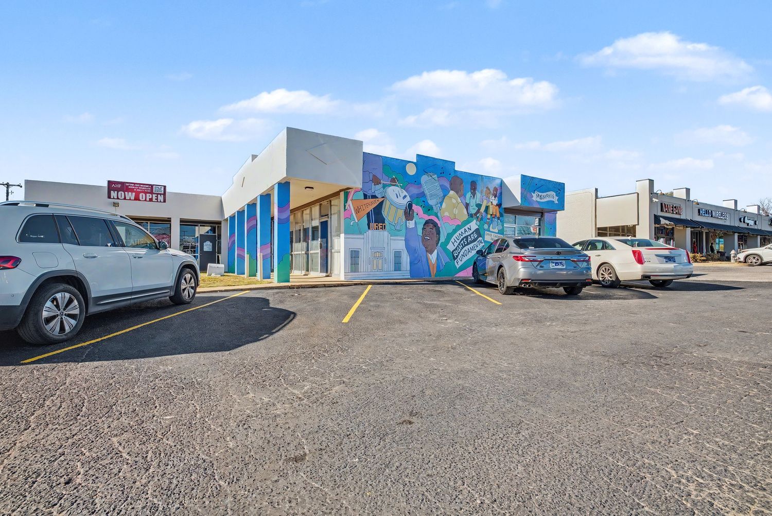 Panoramic view of a commercial strip mall with colorful murals and parked cars in a gravel parking lot.
