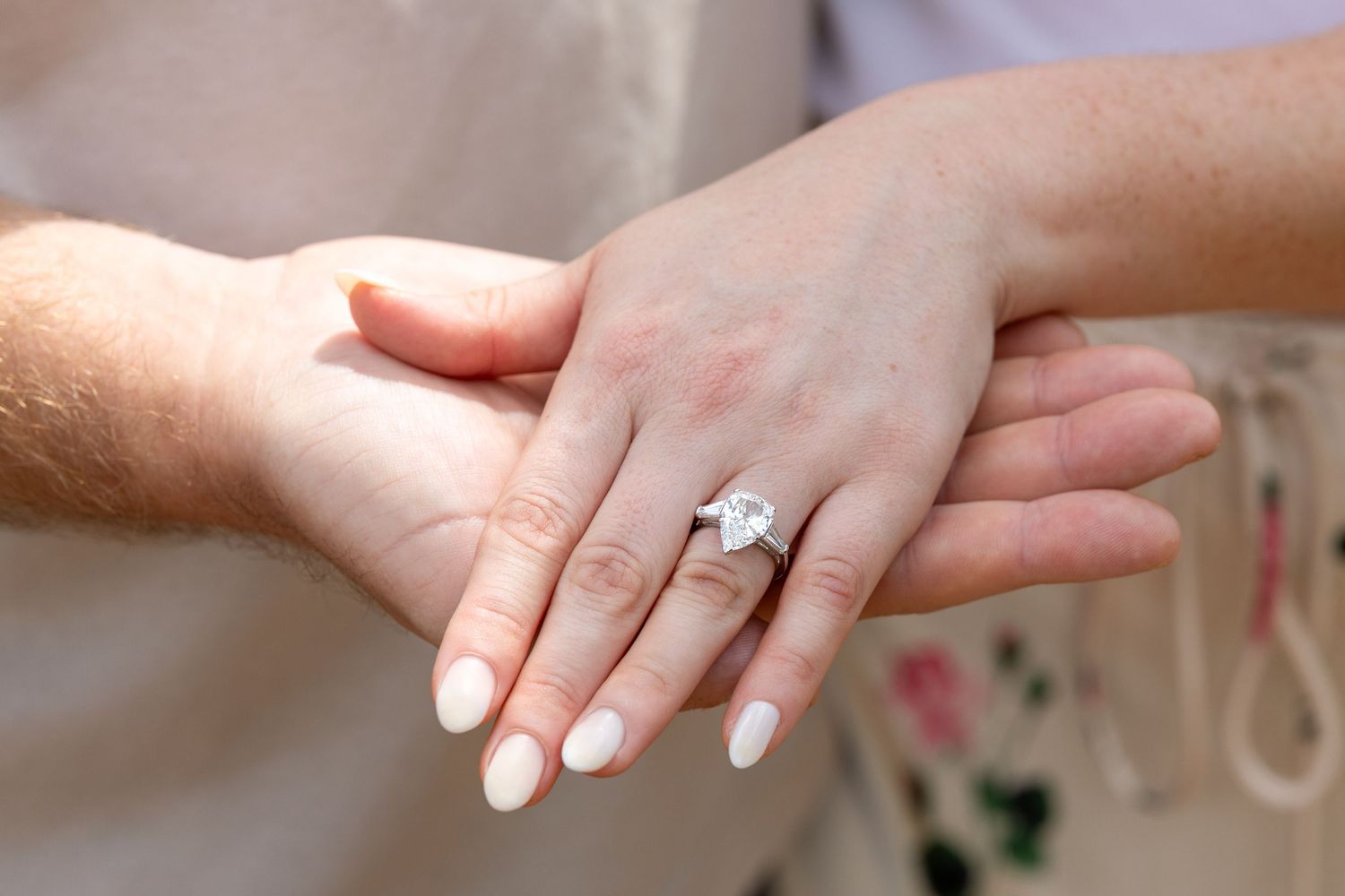 Close-up of a woman's hand wearing a stunning diamond engagement ring with white manicured nails resting on a man's hand.