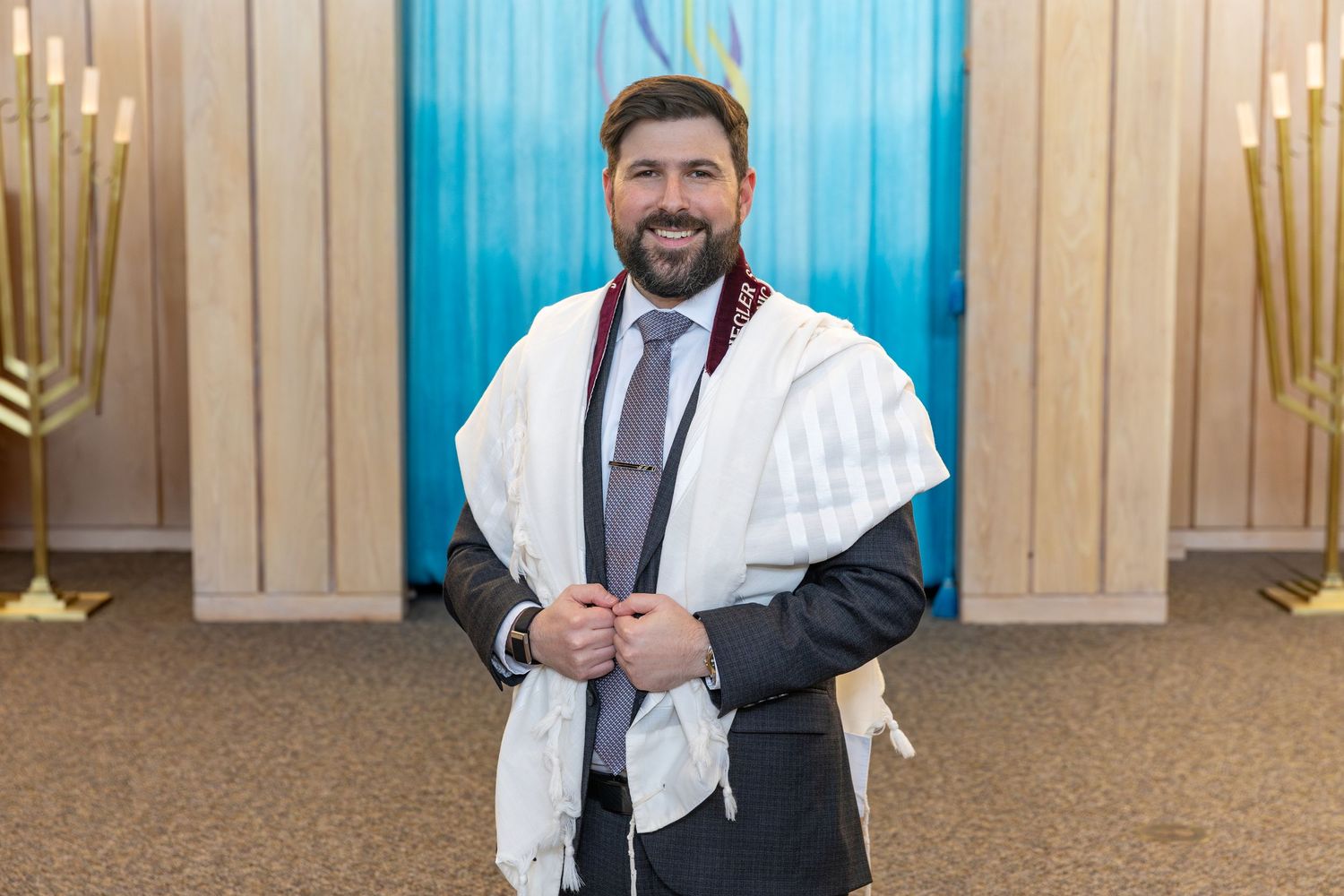Smiling rabbi wearing a white tallit prayer shawl and suit stands in a synagogue with menorahs visible.