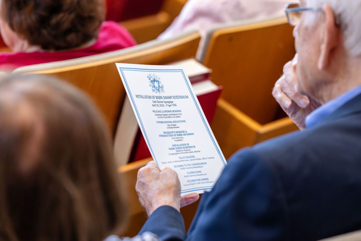 Elderly man and woman sitting in church pews reviewing a printed program during a religious service.