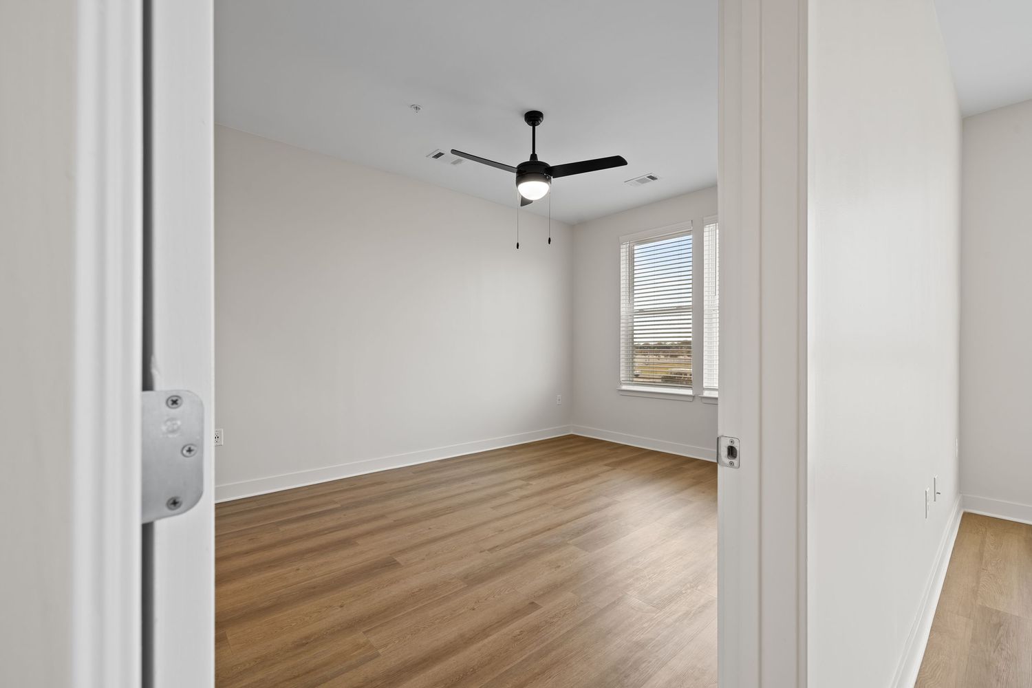 Empty room with hardwood floors, ceiling fan, and large windows offering natural light.