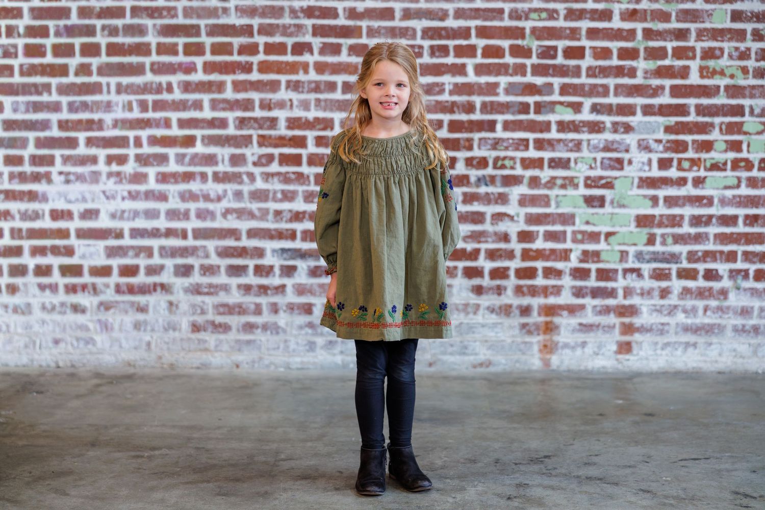 A person wearing a green military-style jacket stands against a red brick wall.
