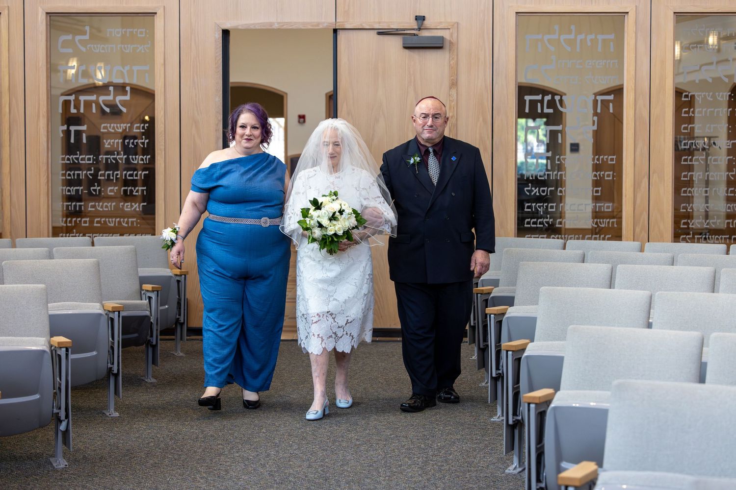 Wedding party members walk through a church hallway lined with wooden cabinets and grey chairs.