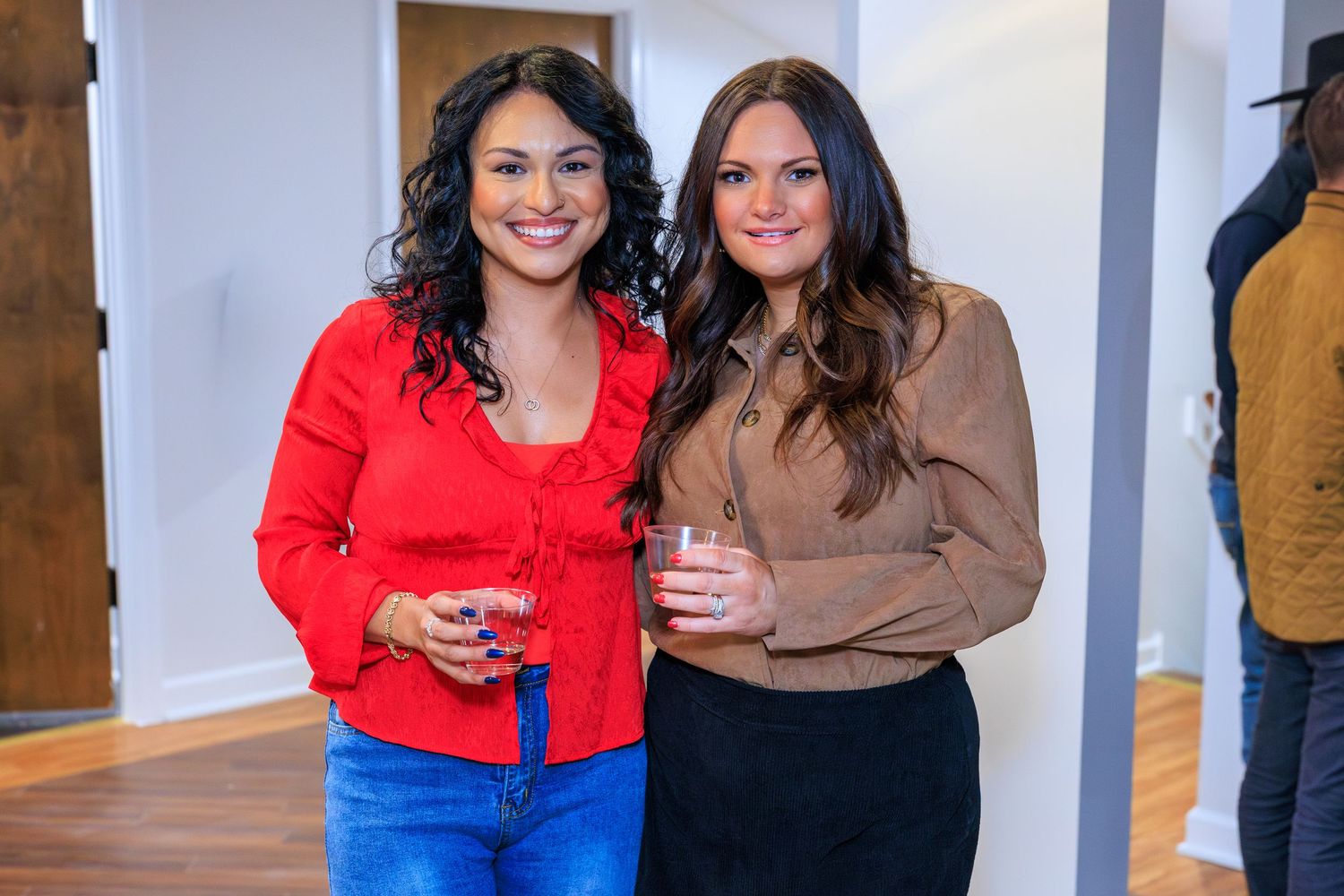 Two friends wearing red and brown sweaters pose together in front of a light colored wall.
