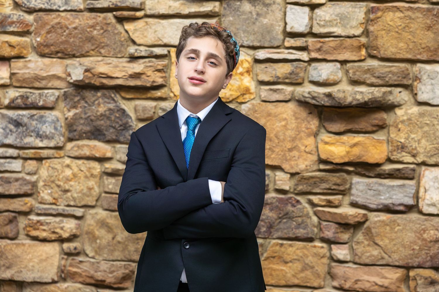 A person in a navy suit and blue tie stands confidently with arms crossed in front of a stone wall.