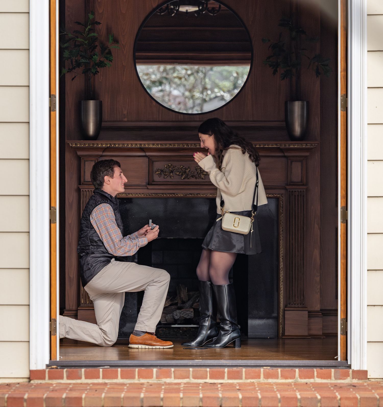 Man on one knee proposing to woman in doorway with fireplace and round mirror backdrop.