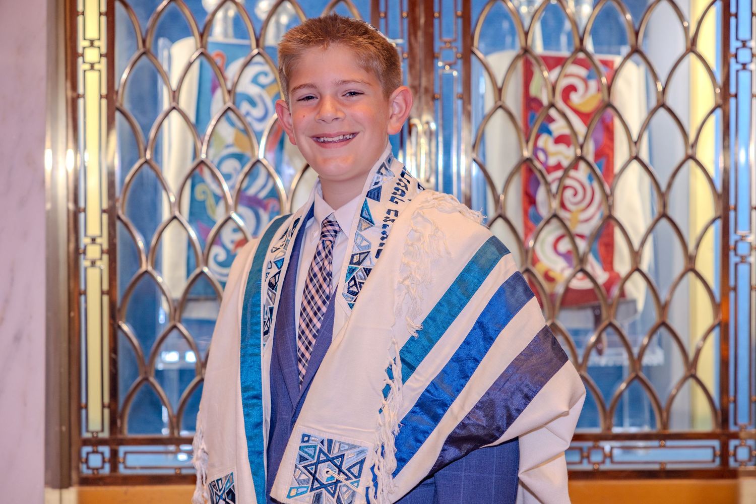 Smiling boy in tallit and blue suit at Bar Mitzvah ceremony in synagogue with stained glass windows.