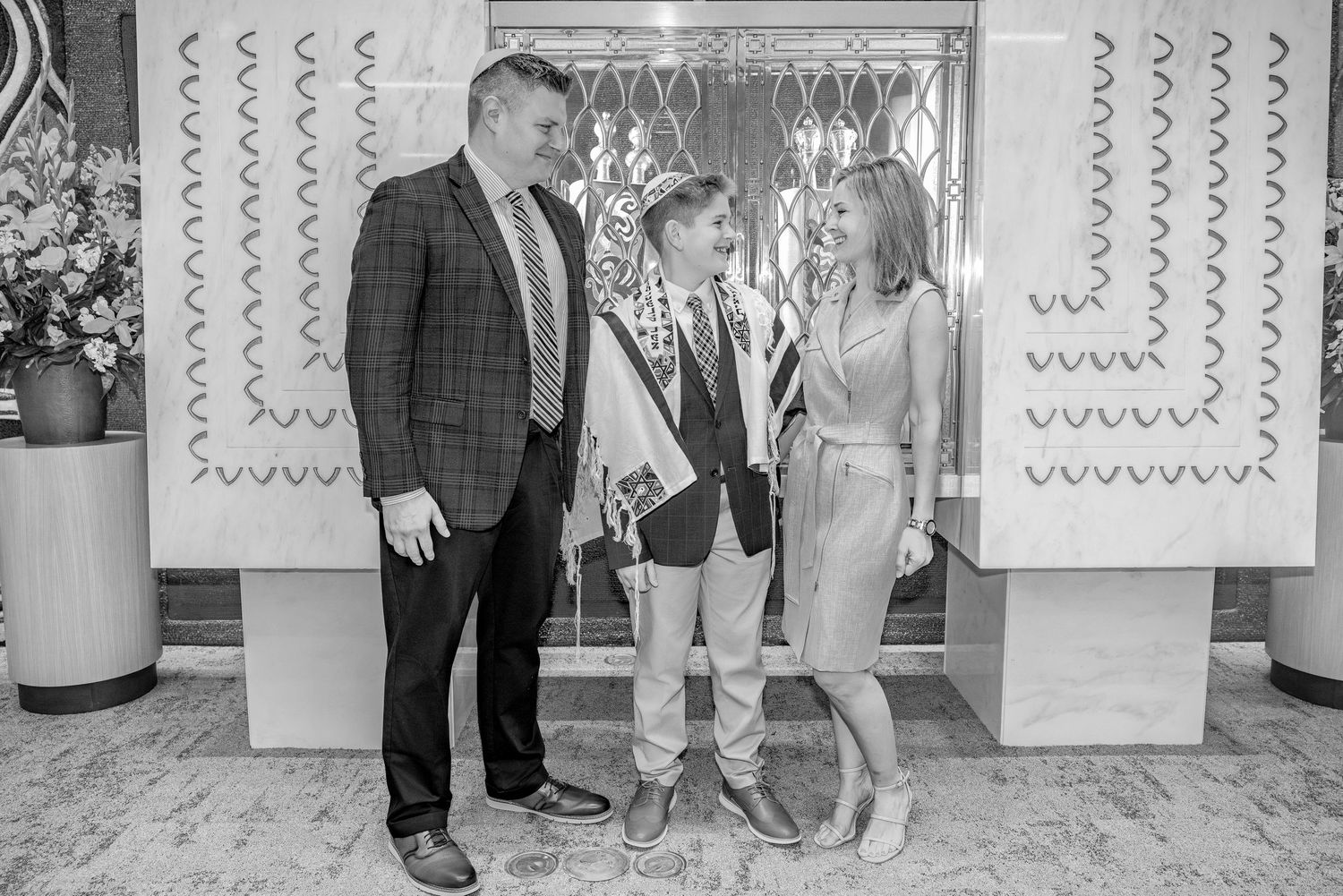 Family of three posing together indoors, boy wearing sash between smiling man in blazer and woman in dress.