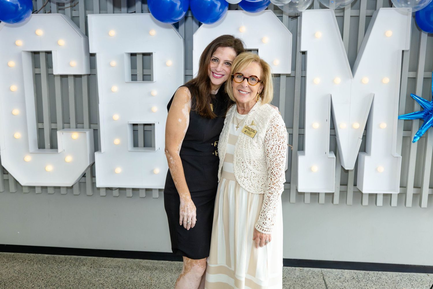 Two people pose together at an event with large white letters and blue balloons in the background.
