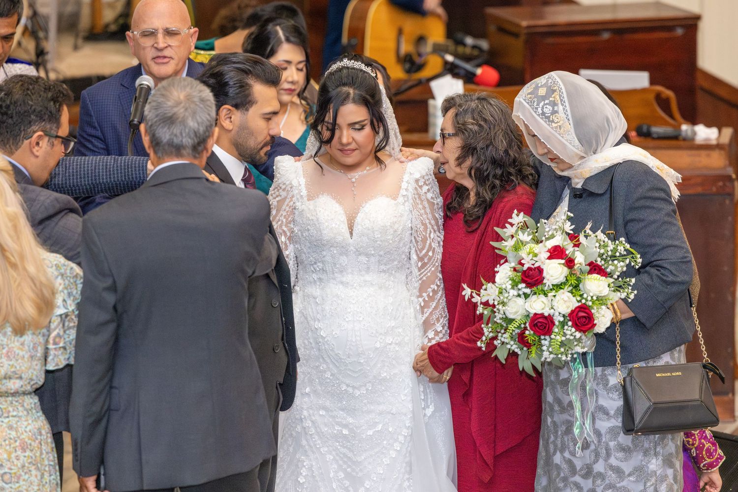 Wedding celebration with guests surrounding couple in formal attire and red floral arrangements.