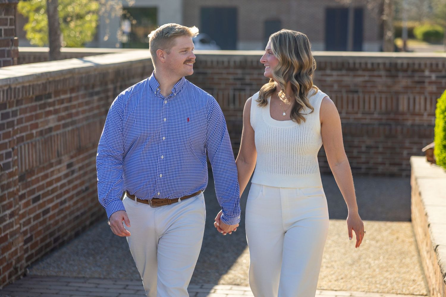A couple walks hand-in-hand smiling at each other near a brick wall on a sunny day.