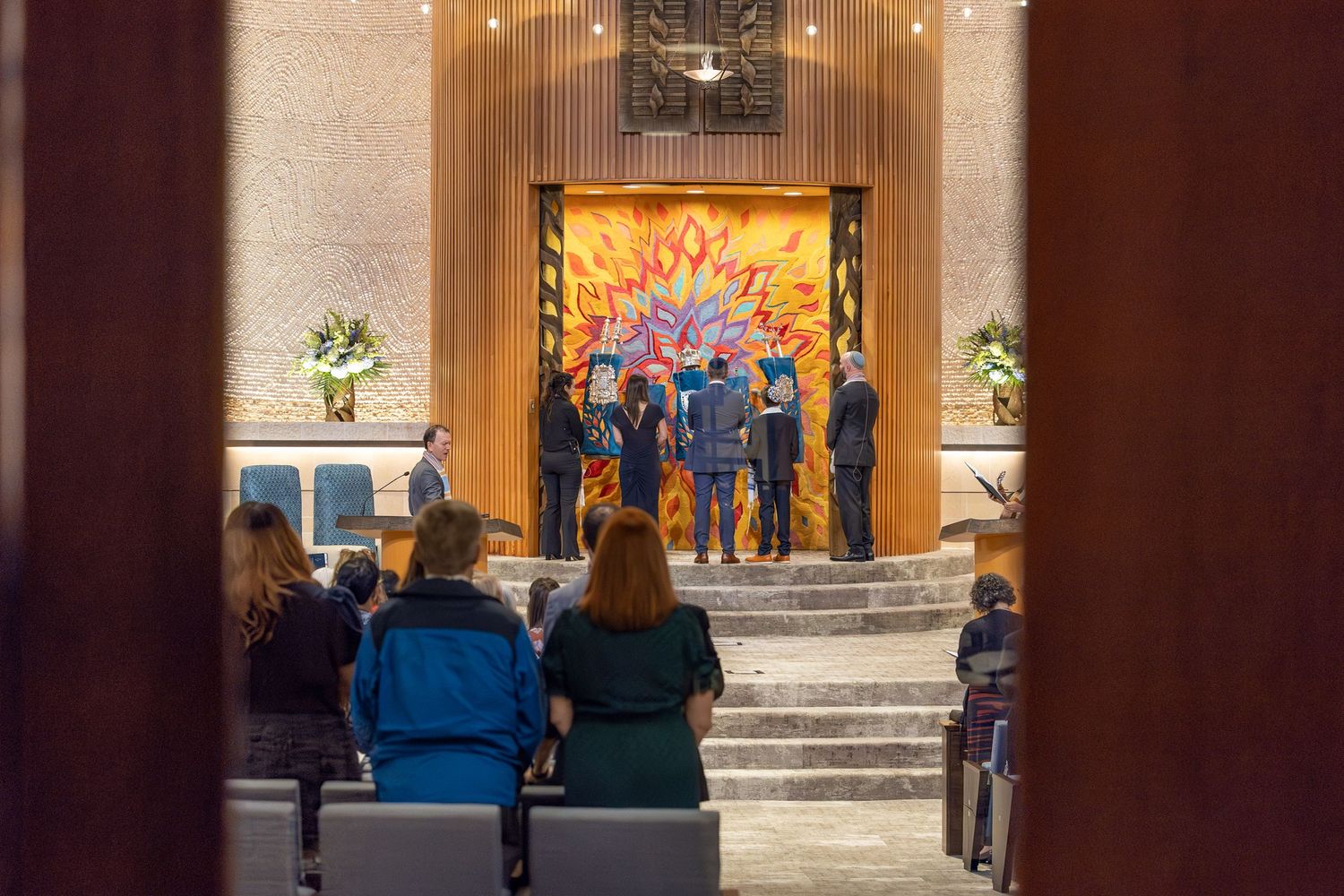 Wedding ceremony in a modern chapel with colorful stained glass altar, guests seated in pews, and wedding party at the front.