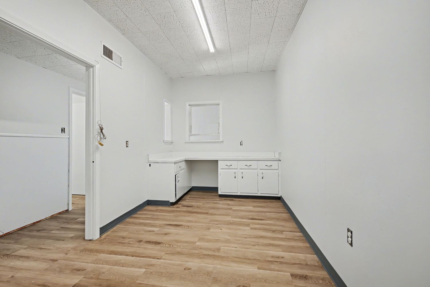 Empty white interior room with hardwood flooring, fluorescent lighting, and baseboards along the walls.