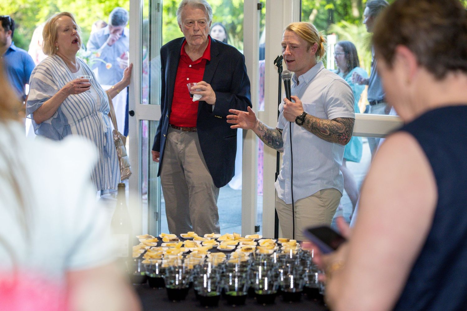 Man with microphone speaks at elegant outdoor event with guests mingling near trays of appetizers.