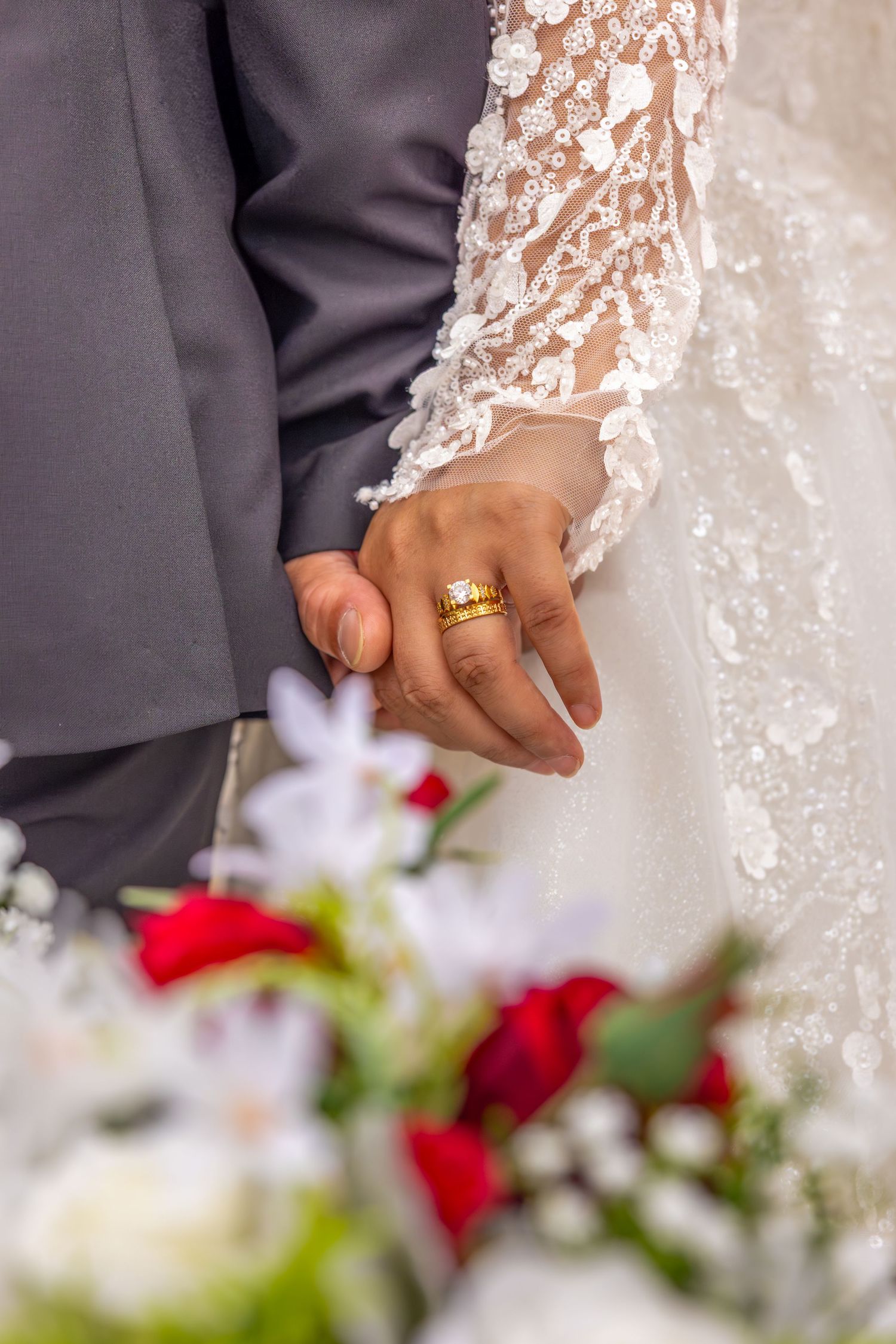 Close-up detail of wedding attire showing intricate lace dress and suit sleeve with red and white bouquet.