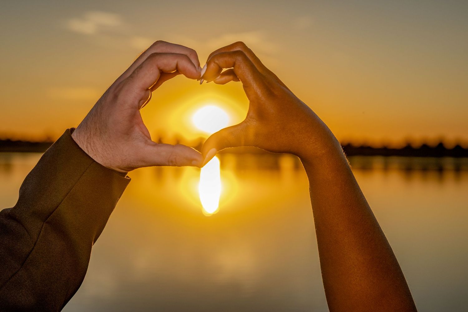 Hands forming a heart shape silhouetted against a golden sunset over water.