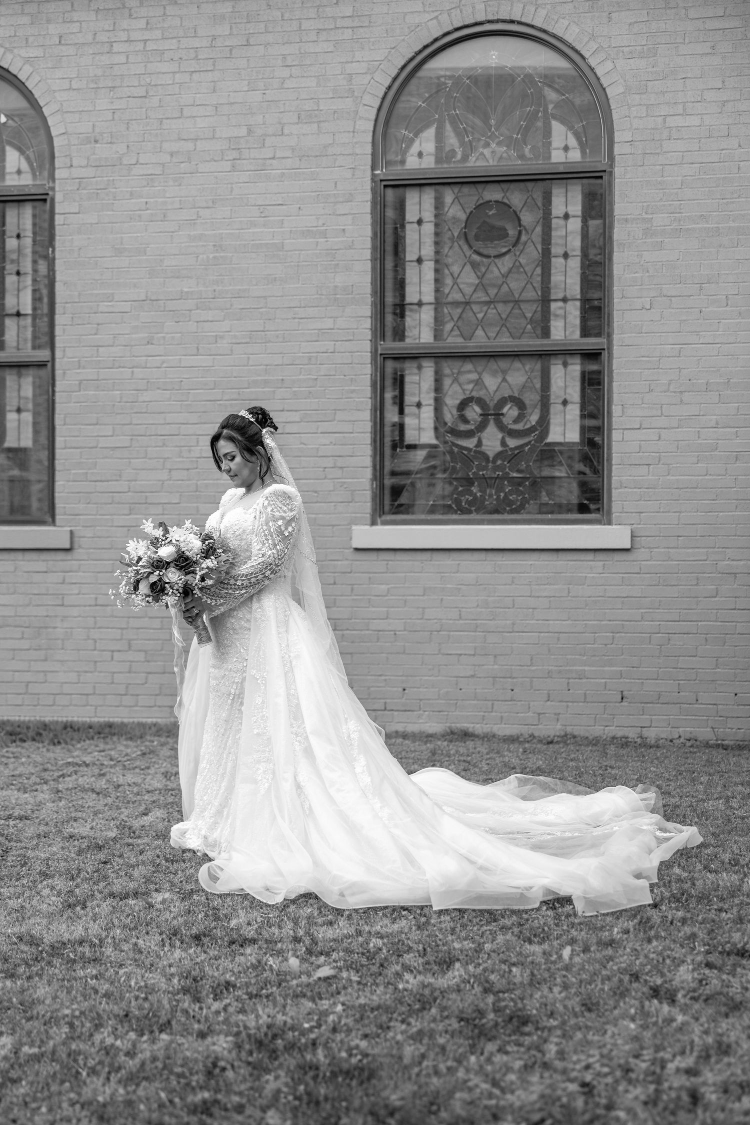 Black and white photo of a bride in a flowing white dress with long train on church grounds.
