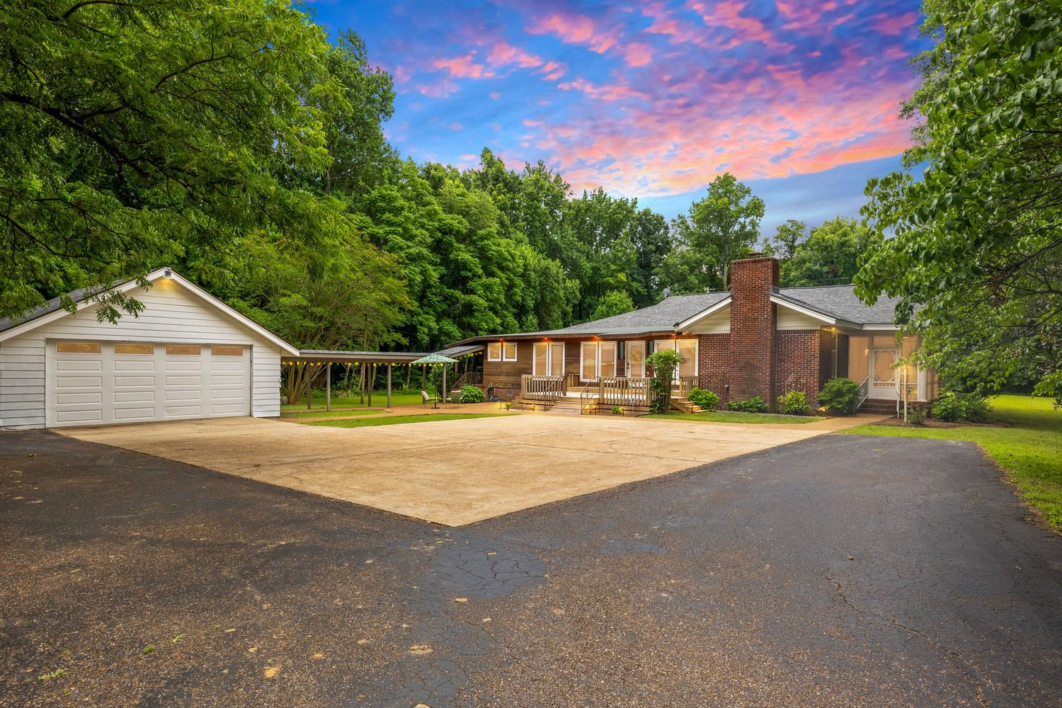 Brick ranch home with detached garage, circular driveway, and lush trees under a vibrant sunset sky.