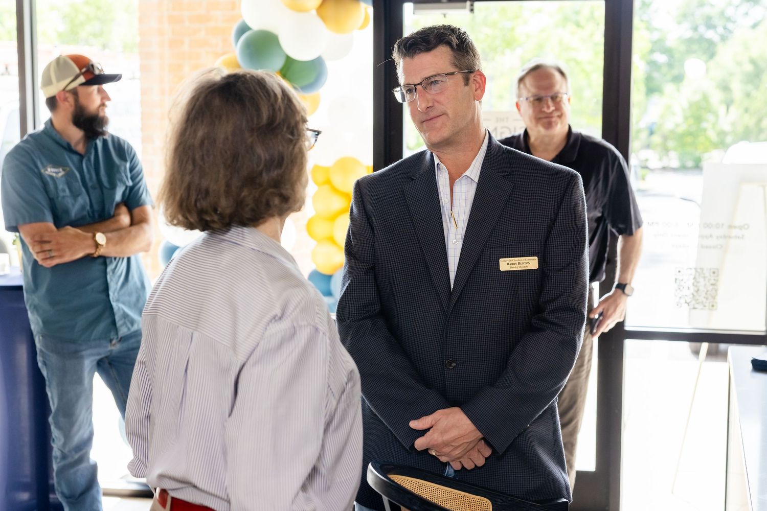 Professional man in suit with name tag conversing with woman at an indoor event with colorful balloons.
