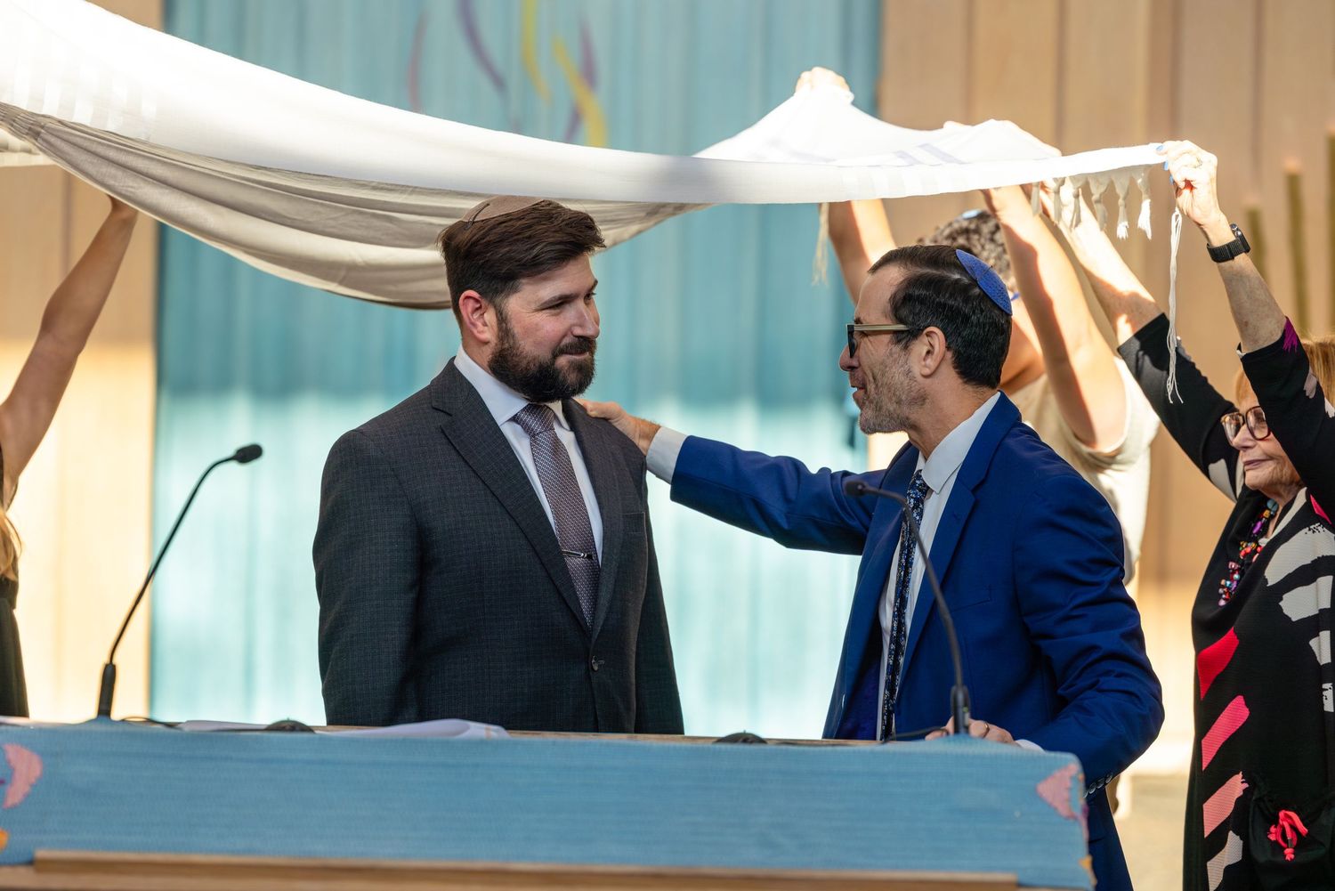 Two men in suits stand under a chuppah during a Jewish wedding ceremony in a synagogue.