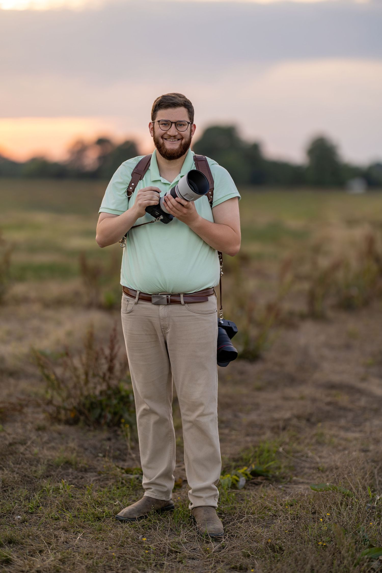 Smiling bearded male photographer holding camera with telephoto lens standing in field at sunset.