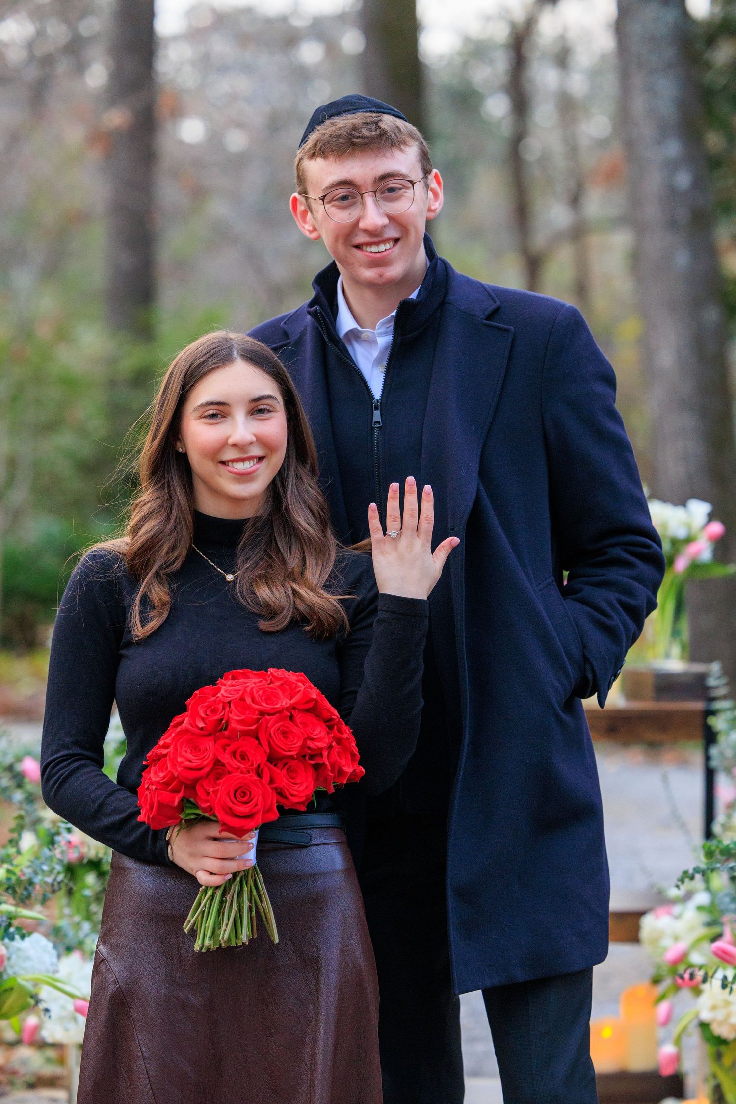 A couple poses outdoors in winter attire with a vibrant bouquet of red roses.