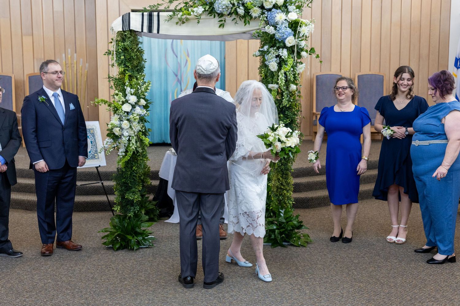 Wedding party gathers in front of a wooden wall decorated with white floral arrangements and greenery.