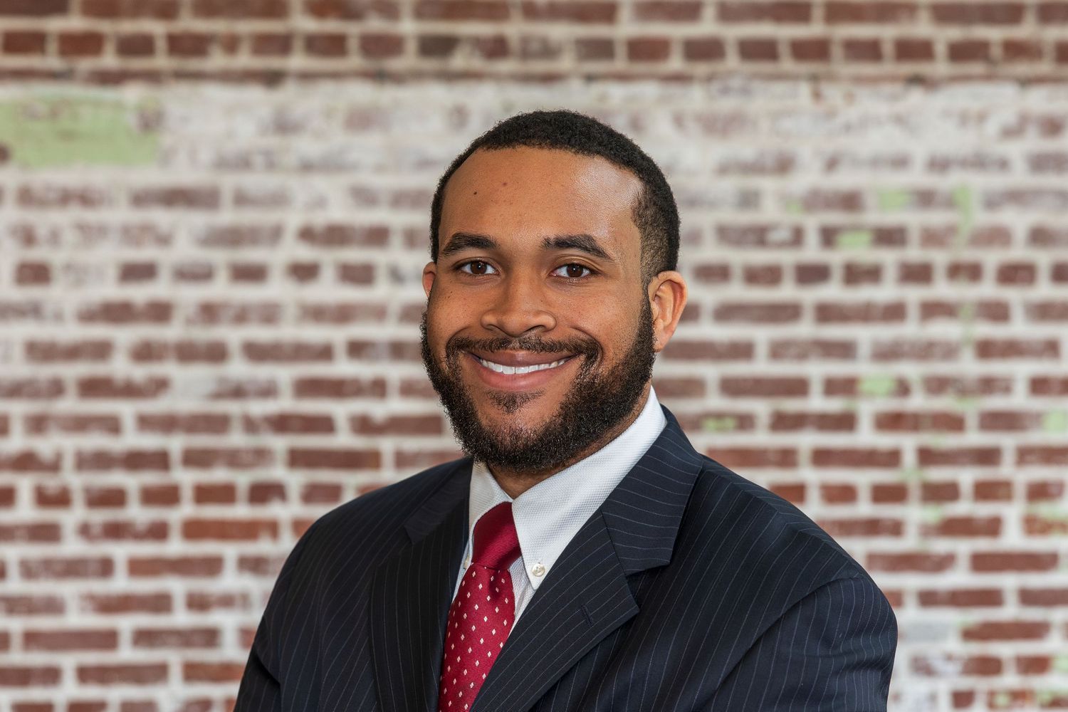 Professional headshot of someone in a dark suit and red tie standing in front of a brick wall.
