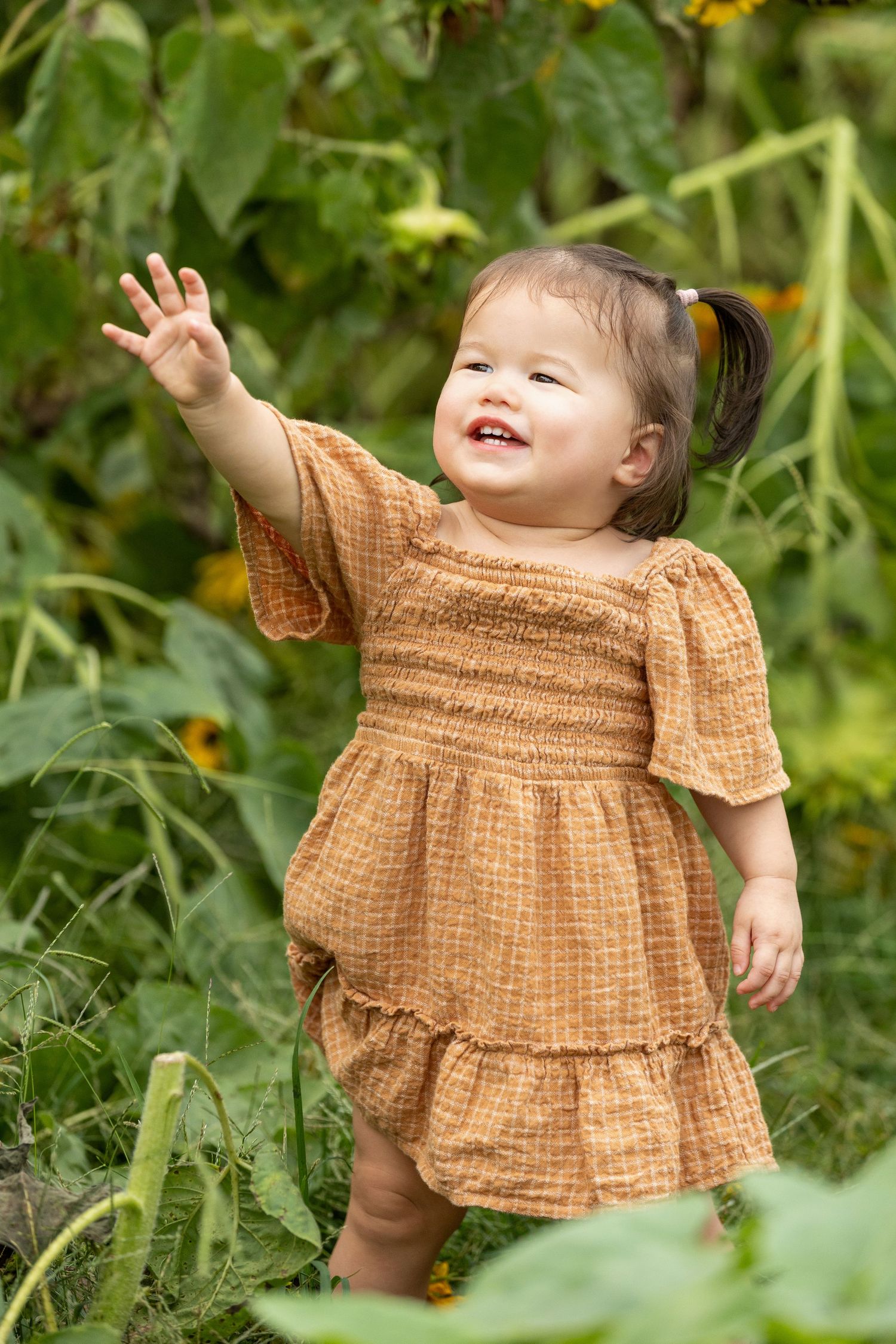 Small child in brown dress playing joyfully among garden greenery.