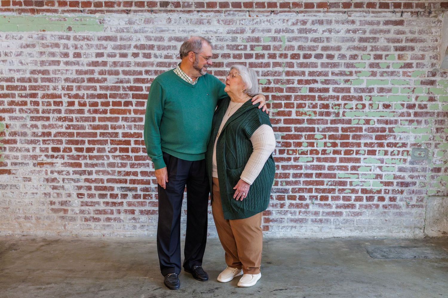 Two people in casual attire stand together against a weathered brick wall.