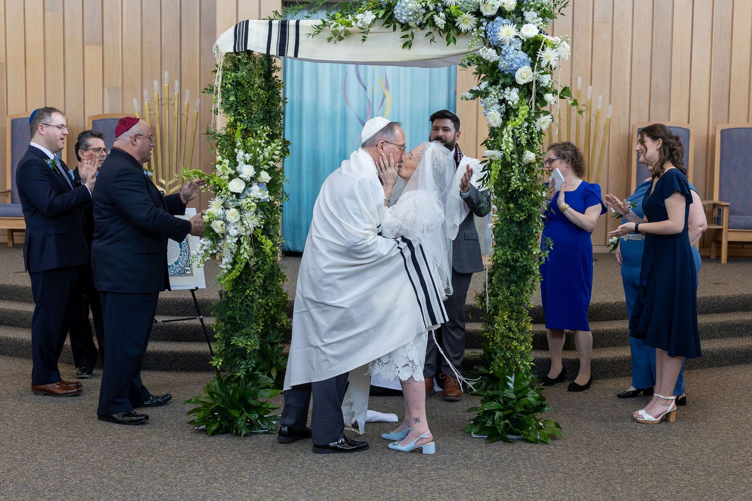 Jewish wedding ceremony under floral chuppah with couple kissing surrounded by family in synagogue.