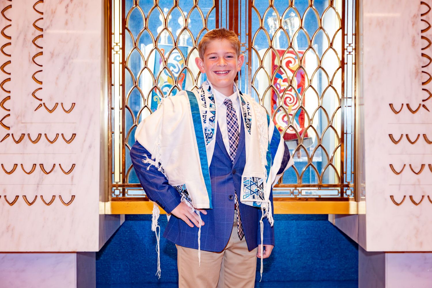 Young boy in blue blazer and tallit smiling at his Bar Mitzvah ceremony in a synagogue.