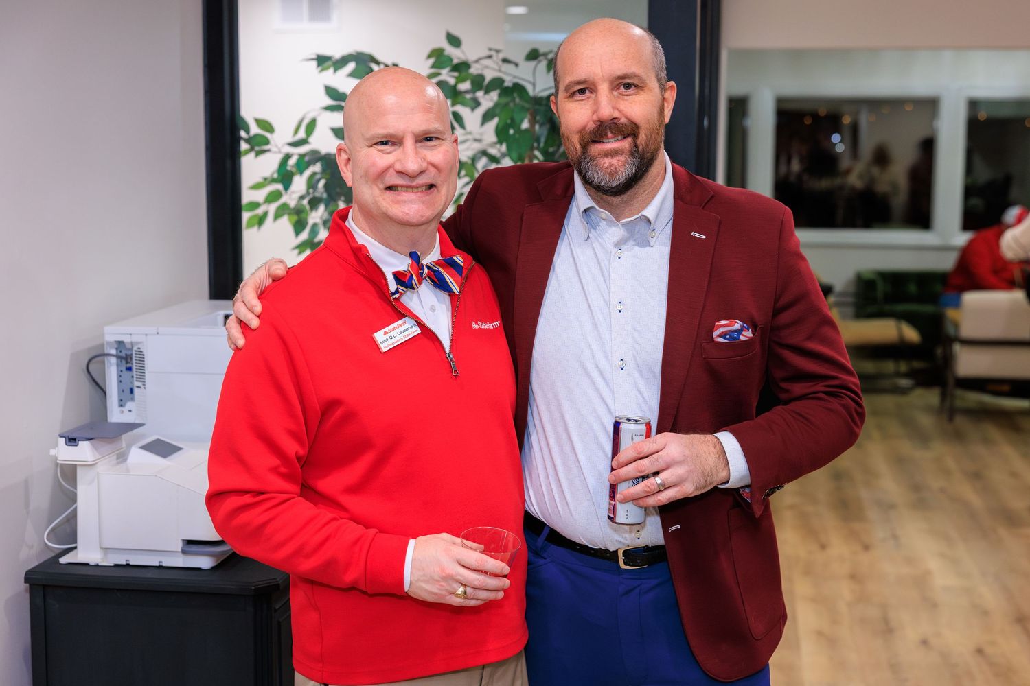 Two people pose together at an event wearing red and burgundy jackets while holding beverages.